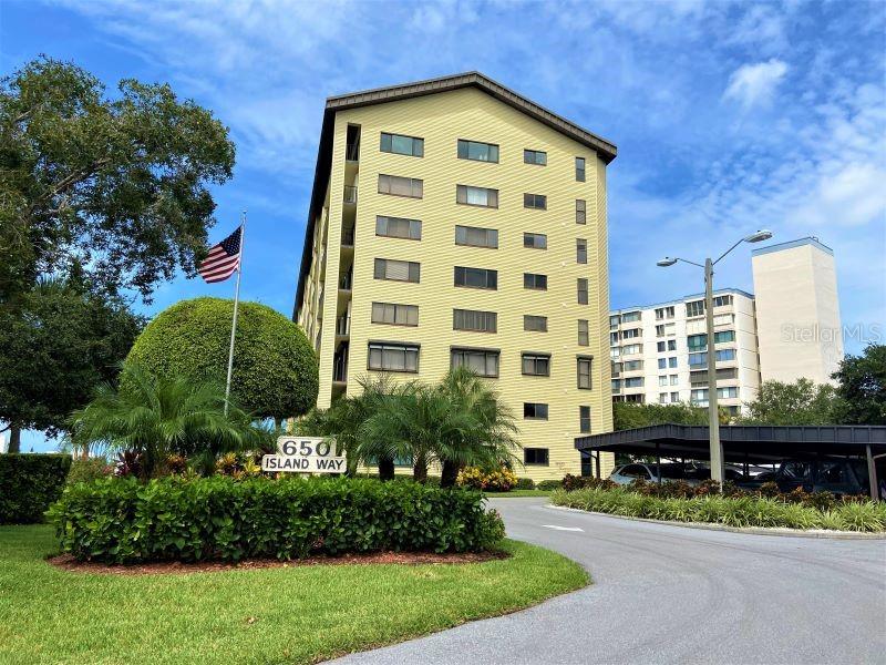 A yellow multi-story apartment building stands behind a landscaped lawn and a covered parking area — 650 ISLAND WAY UNIT 708, CLEARWATER BEACH, FL