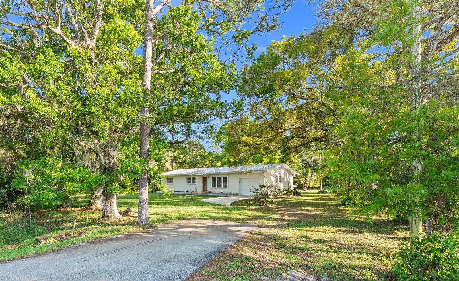 A single-story house with a white exterior and metal roof sits among many large trees — 11541 Okeechobee Road, Fort Pierce FL 34945, FORT PIERCE, FL
