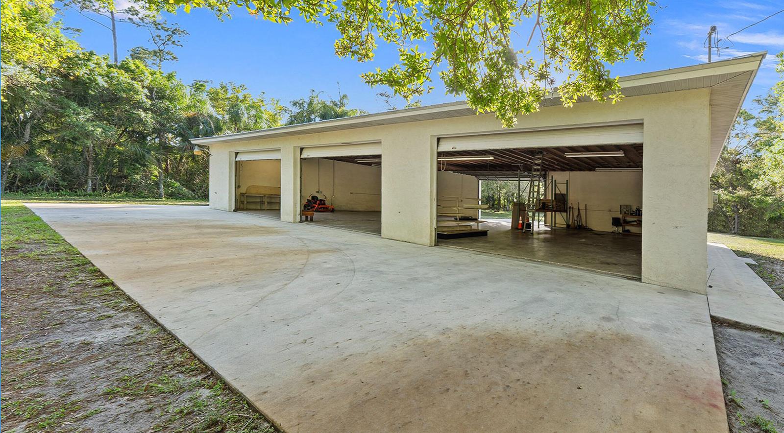 A detached three-bay garage with a concrete driveway sits in a wooded outdoor setting — 11541 Okeechobee Road, Fort Pierce FL 34945, FORT PIERCE, FL