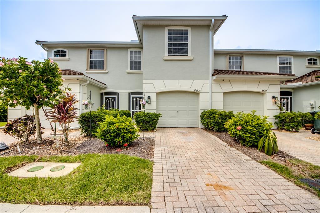 A two-story townhouse exterior with a paved driveway, two garage doors, and landscaped front yard — 8668 ATHENA COURT, LEHIGH ACRES, FL
