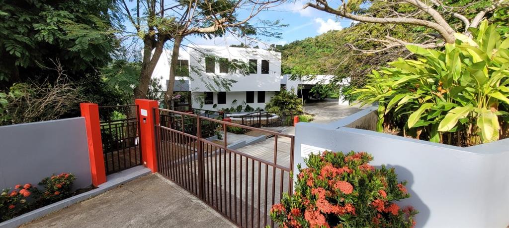 A white two-story house is visible behind a metal gate and a gray wall — PR 992 KM3.4 3 ST VILLAS DE LUQUILLO UNIT D18, LUQUILLO, PR