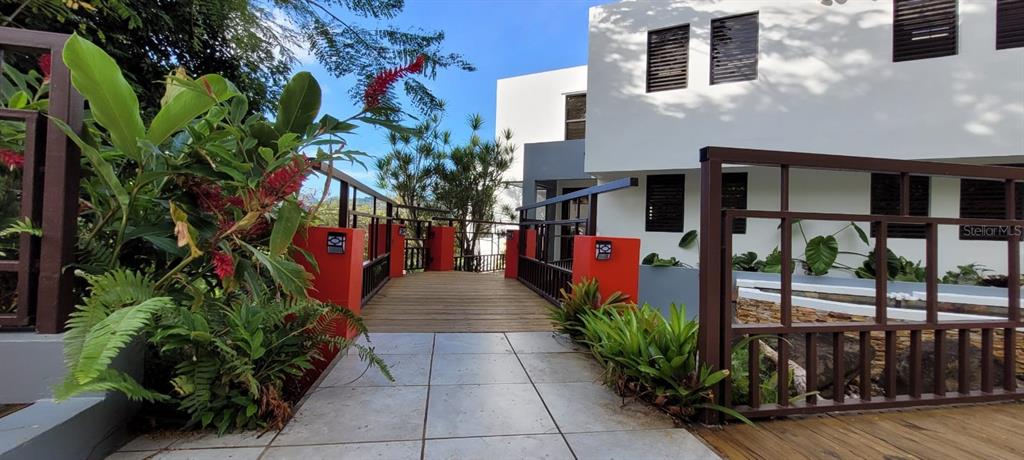 A wooden bridge leads to the entrance of a modern white house with red accents — PR 992 KM3.4 3 ST VILLAS DE LUQUILLO UNIT D18, LUQUILLO, PR