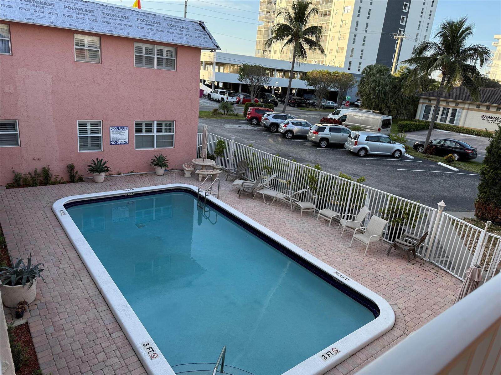 An outdoor swimming pool with a white fence is situated beside a pink building — 1450 N Riverside Drive # 208, Pompano Beach FL 33062, POMPANO BEACH, FL