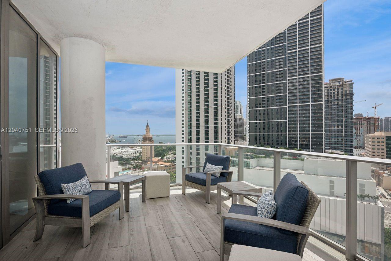 A balcony with wood-look tile flooring features three armchairs and two small tables overlooking city buildings — 851 NE 1st Ave # 1711, Miami FL 33132, MIAMI, FL