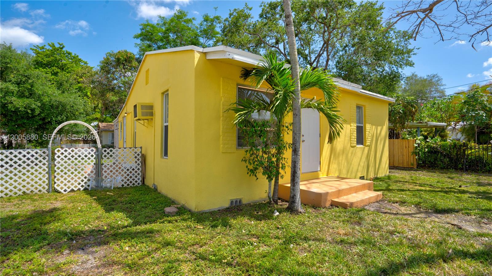 A bright yellow, single-story house features a small front porch and a white lattice fence — 1085 NE 128th St, North Miami FL 33161, NORTH MIAMI, FL