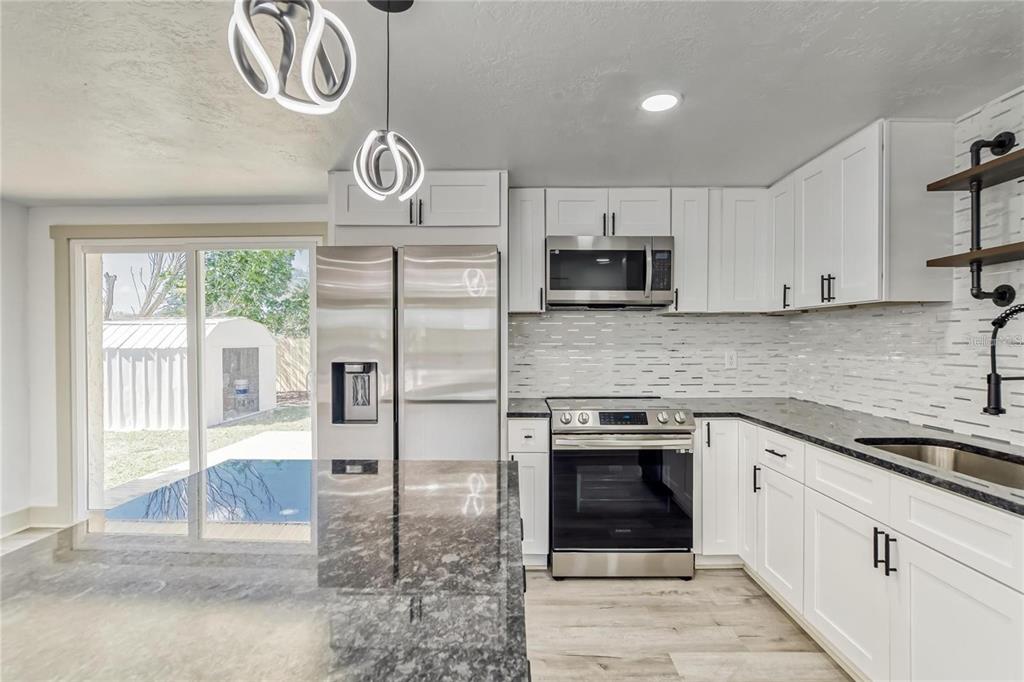 A modern kitchen features white cabinets, stainless steel appliances, dark granite countertops, and sliding doors — 1300 MELONWOOD AVENUE, CLEARWATER, FL