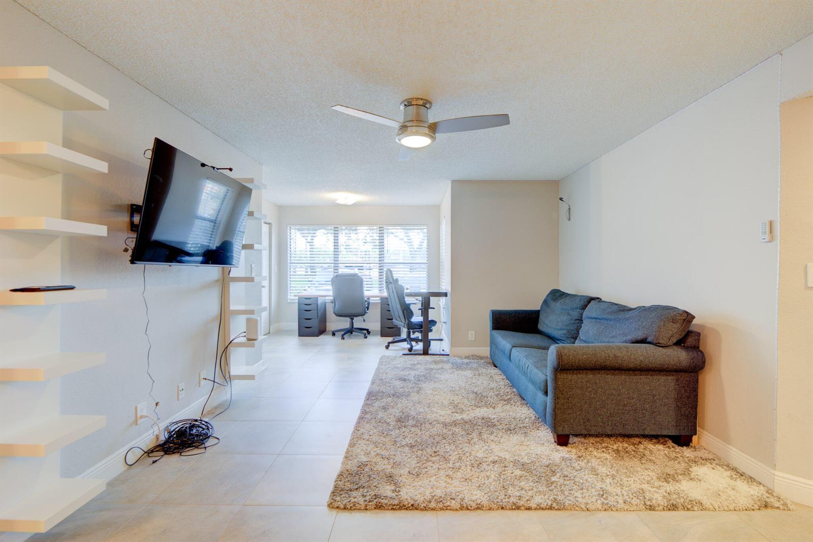 A living room features a blue sofa on a rug, a wall-mounted television, and desks — 2900 S University Drive # 9104, Davie FL 33328, DAVIE, FL