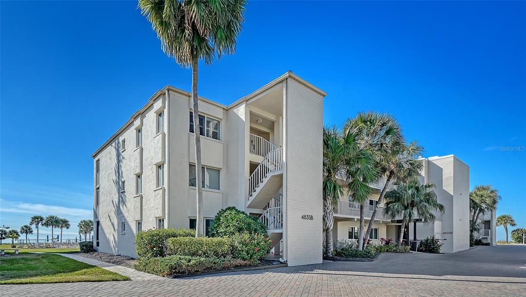 A multi-story beige apartment building features exterior staircases and palm trees under a blue sky — 4835 GULF OF MEXICO DRIVE UNIT 106, LONGBOAT KEY, FL