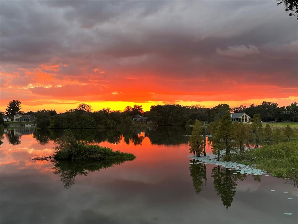 A vibrant sunset reflects over a lake with trees and houses along the shoreline — 858 HANOVER WAY, LAKELAND, FL