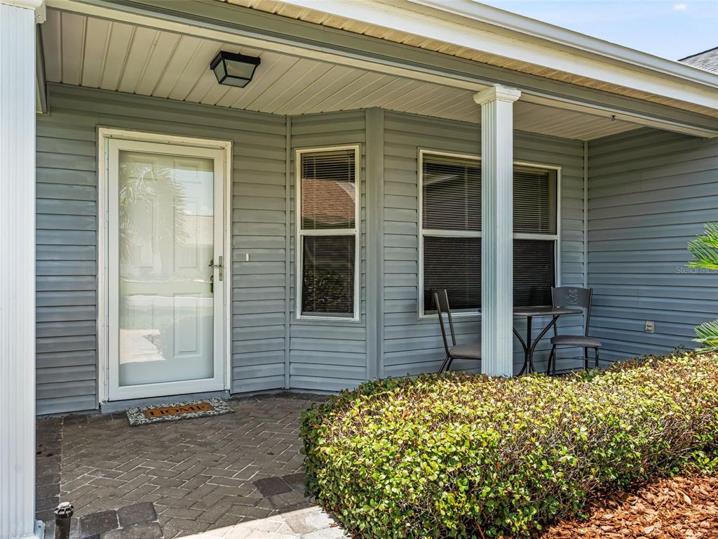 A covered front porch features gray vinyl siding, a brick paver floor, and two chairs — 1506 ROCHA LANE, THE VILLAGES, FL