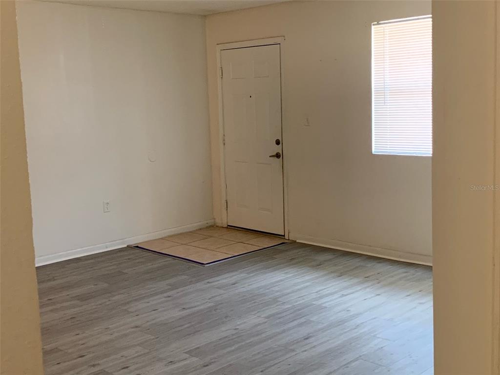 An empty room features light gray wood-look flooring, white walls, a white door, and tiled entryway — 12403 HIBISCUS OAK PLACE UNIT 202, TAMPA, FL