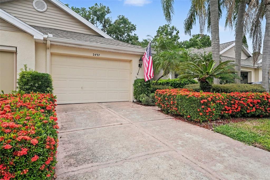 A beige house exterior features a garage, a concrete driveway, and vibrant red flowering bushes — 2437 NANTUCKET HARBOR LOOP UNIT 2437, SUN CITY CENTER, FL