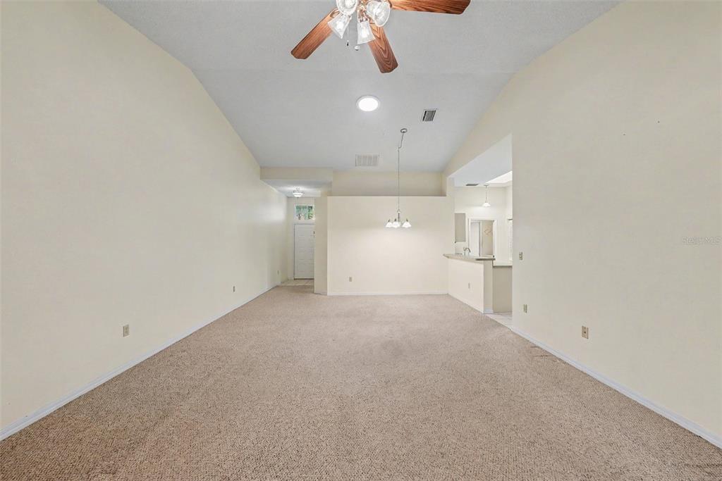 An empty living room with beige carpet, vaulted ceilings, and an adjacent kitchen area — 2437 NANTUCKET HARBOR LOOP UNIT 2437, SUN CITY CENTER, FL