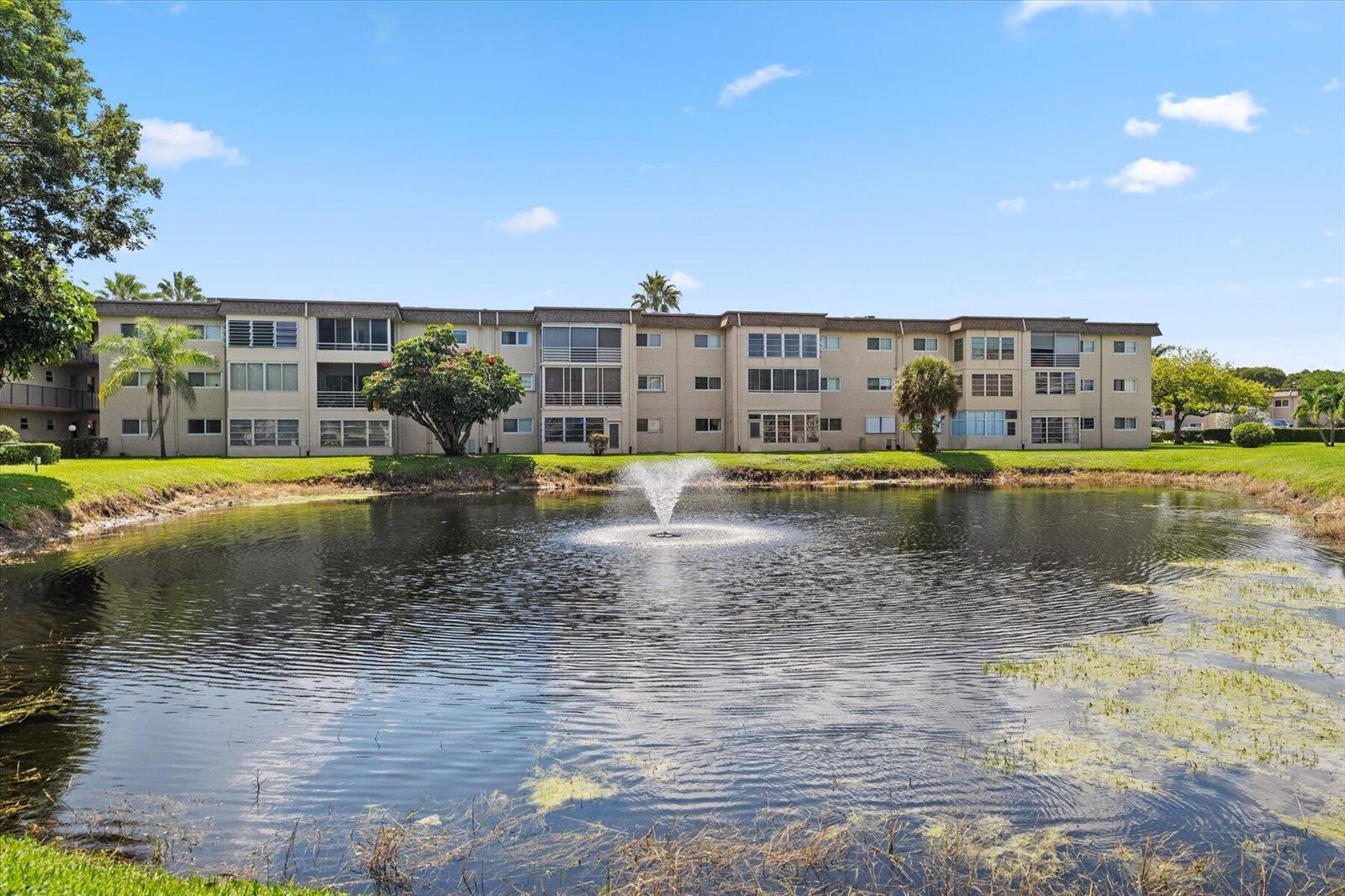A multi-story apartment building sits behind a pond with a central water fountain — 719 Lori Drive # 204, Palm Springs FL 33461, PALM SPRINGS, FL
