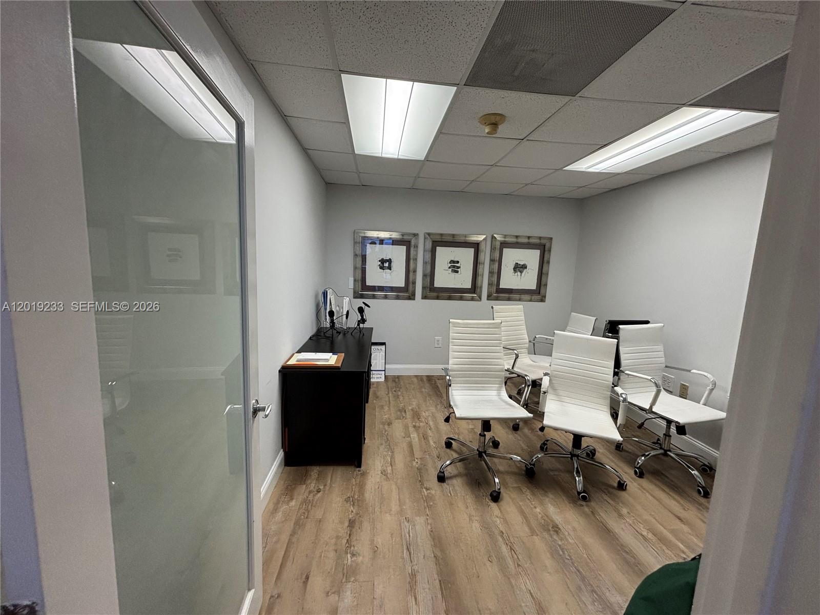 An office room features wood-look flooring, white walls, a black console table, and four chairs — NORTH BAY VILLAGE, FL