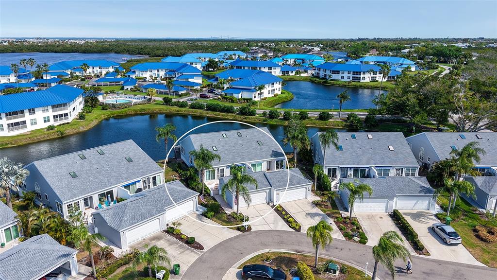 An aerial view shows a residential townhouse complex with blue roofs surrounding a central pond — 1005 PELICAN COURT, BRADENTON, FL