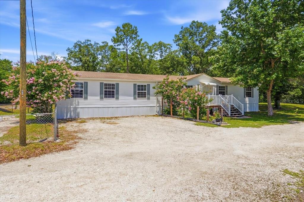 A single-story manufactured home with white siding sits behind a gravel driveway and trees — 10626 PATHFINDER TRAIL, LAKELAND, FL