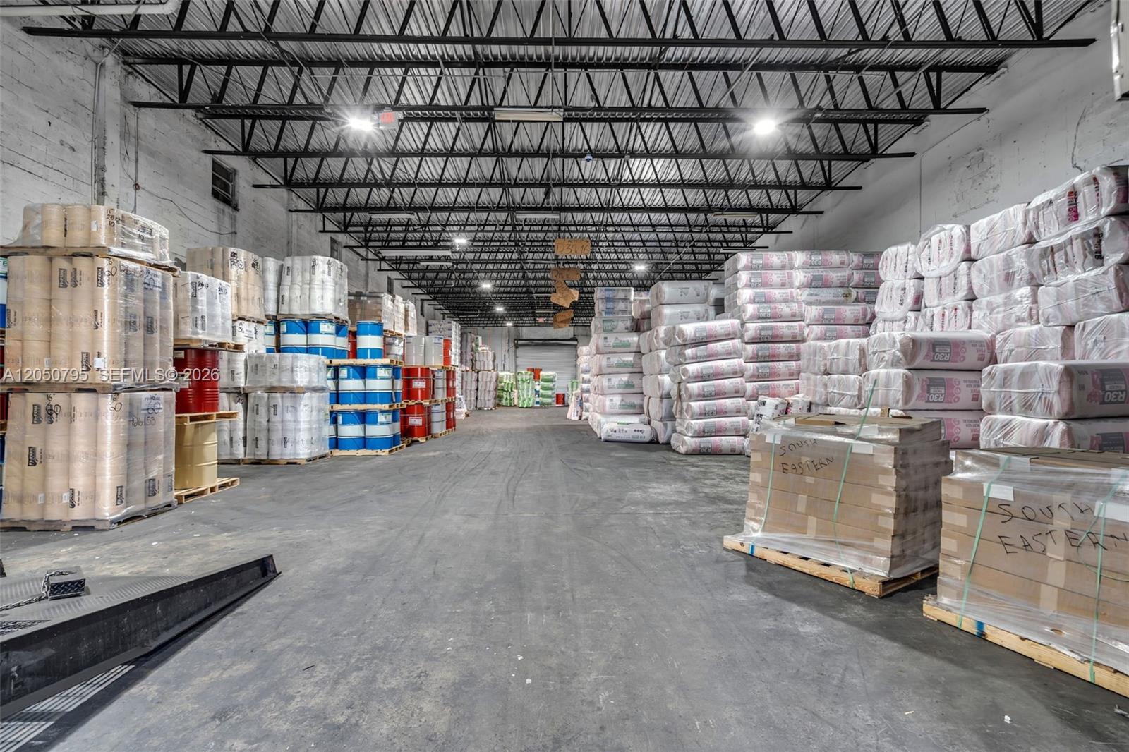 A large warehouse interior features high ceilings with exposed trusses and stacks of palletized inventory — 7440 NW 66th St, Miami FL 33166, MIAMI, FL