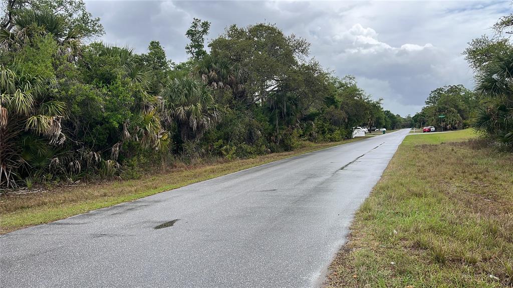 A paved road stretches into the distance, flanked by dense trees and grassy shoulders — 15010 ELWOOD AVENUE, PORT CHARLOTTE, FL