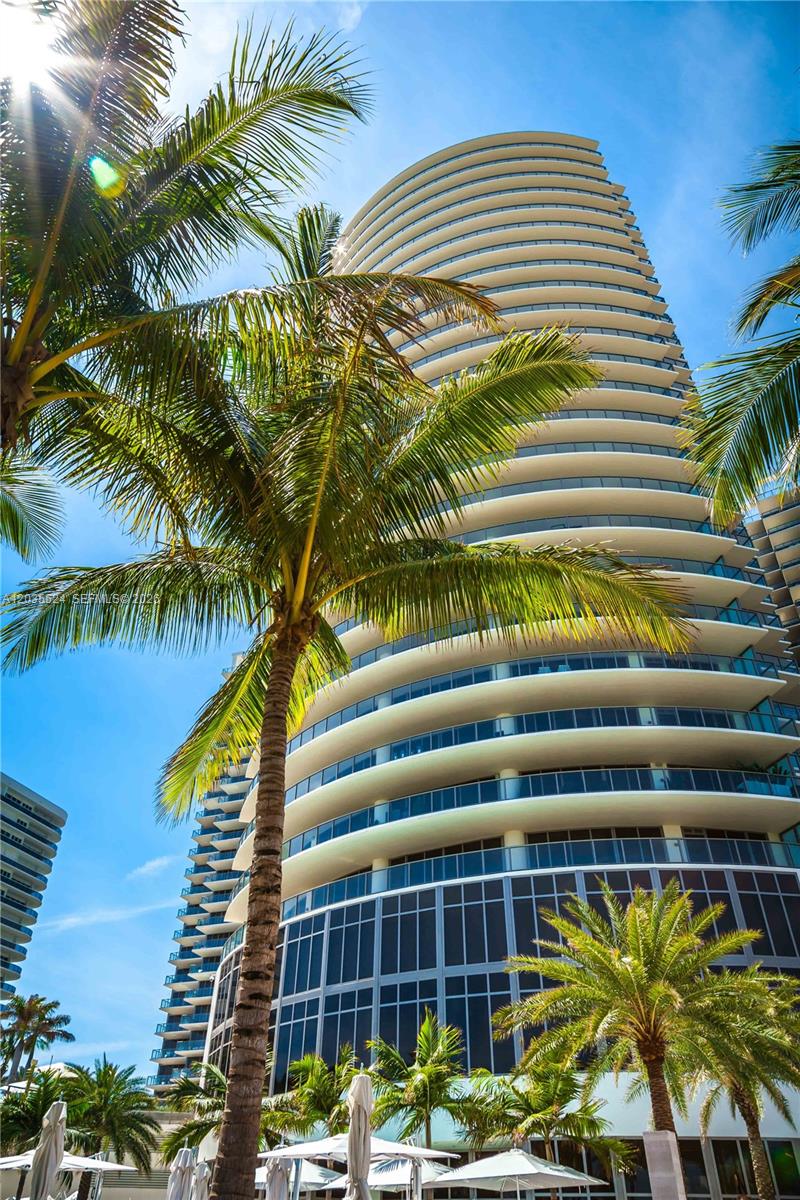 A low-angle view of a modern high-rise building with curved balconies and surrounding palm trees — 9703 Collins Ave, Bal Harbour FL 33154, BAL HARBOUR, FL