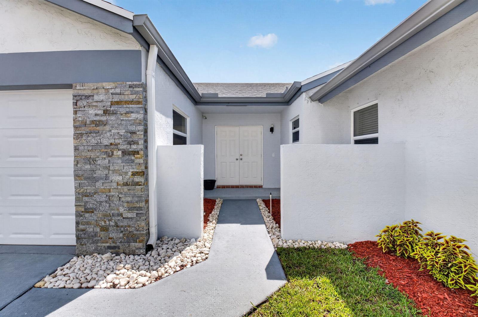 A white house exterior features a stone-accented garage, a central double-door entrance, and landscaping — 21070 Woodspring Avenue, Boca Raton FL 33428, BOCA RATON, FL
