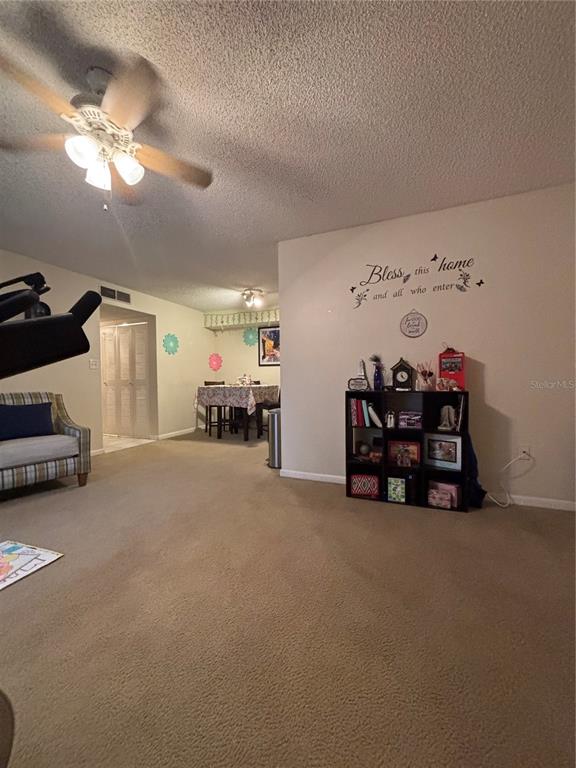 A living area with beige carpet, a ceiling fan, and a bookshelf against a wall — 2690 CORAL LANDINGS BOULEVARD UNIT 127, PALM HARBOR, FL