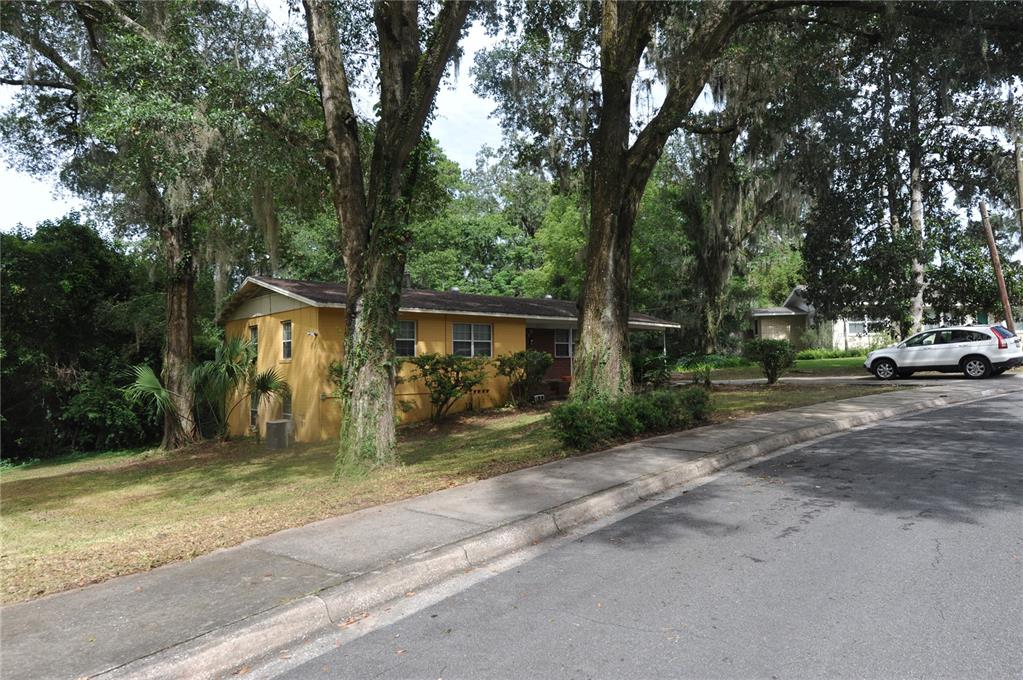 A yellow single-story house sits on a grassy lot with large trees and a sidewalk — 1410 NW 7TH ROAD, GAINESVILLE, FL