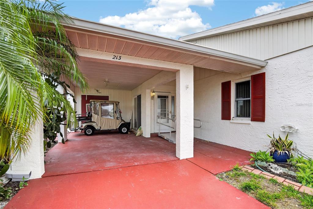 A covered carport with a red concrete floor leads to a house entrance with shutters — 213 GLENELLEN PLACE, SUN CITY CENTER, FL