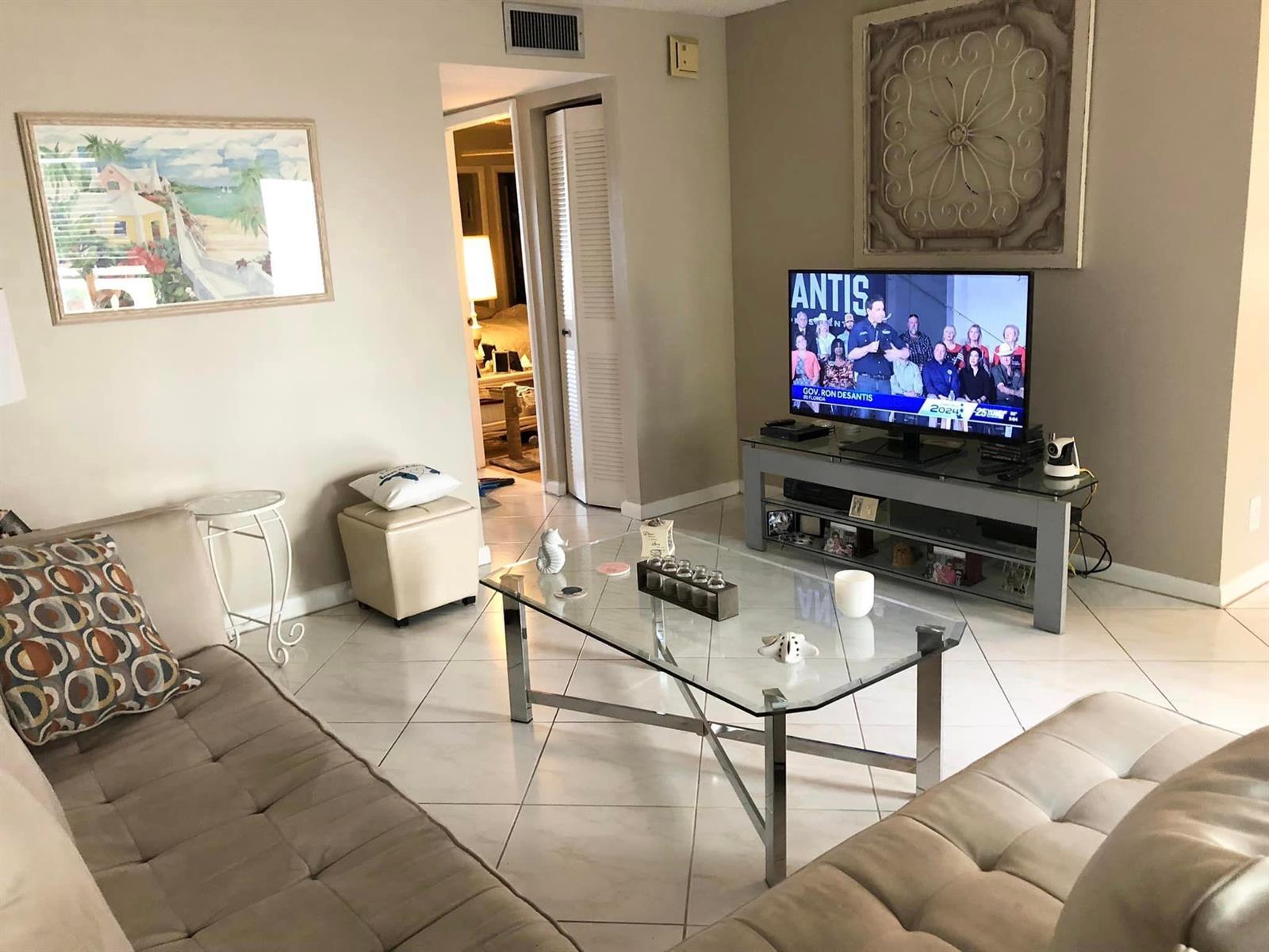 A living room features a glass coffee table, a television on a stand, and tile flooring — 499 Piedmont K, Delray Beach FL 33484, DELRAY BEACH, FL