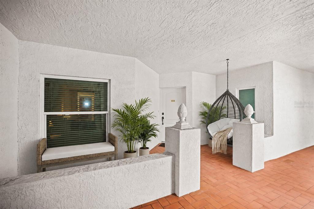 A covered patio features white stucco walls, terracotta tile flooring, a hanging chair, and plants — 200 121ST AVENUE UNIT 103, TREASURE ISLAND, FL