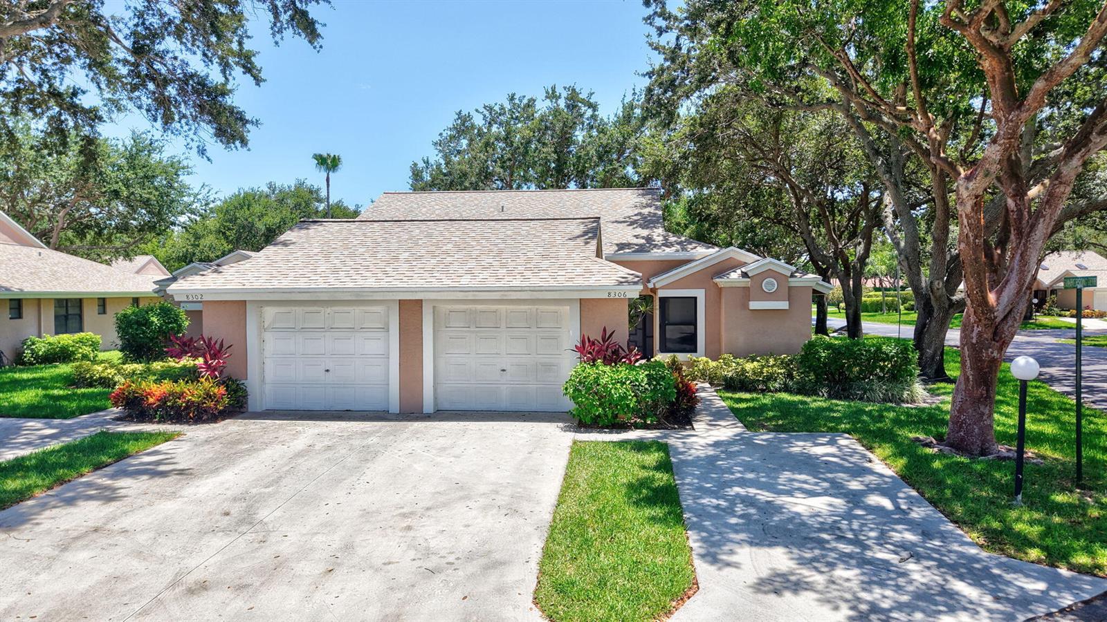 A tan duplex home features two white garage doors and a concrete driveway under trees — 8306 Summersong Terrace, Boca Raton FL 33496, BOCA RATON, FL