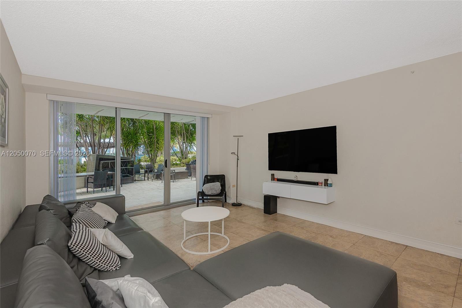 A living room features a gray sectional sofa, a wall-mounted television, and sliding glass doors — 1500 Bay Rd # 116S, Miami Beach FL 33139, MIAMI BEACH, FL