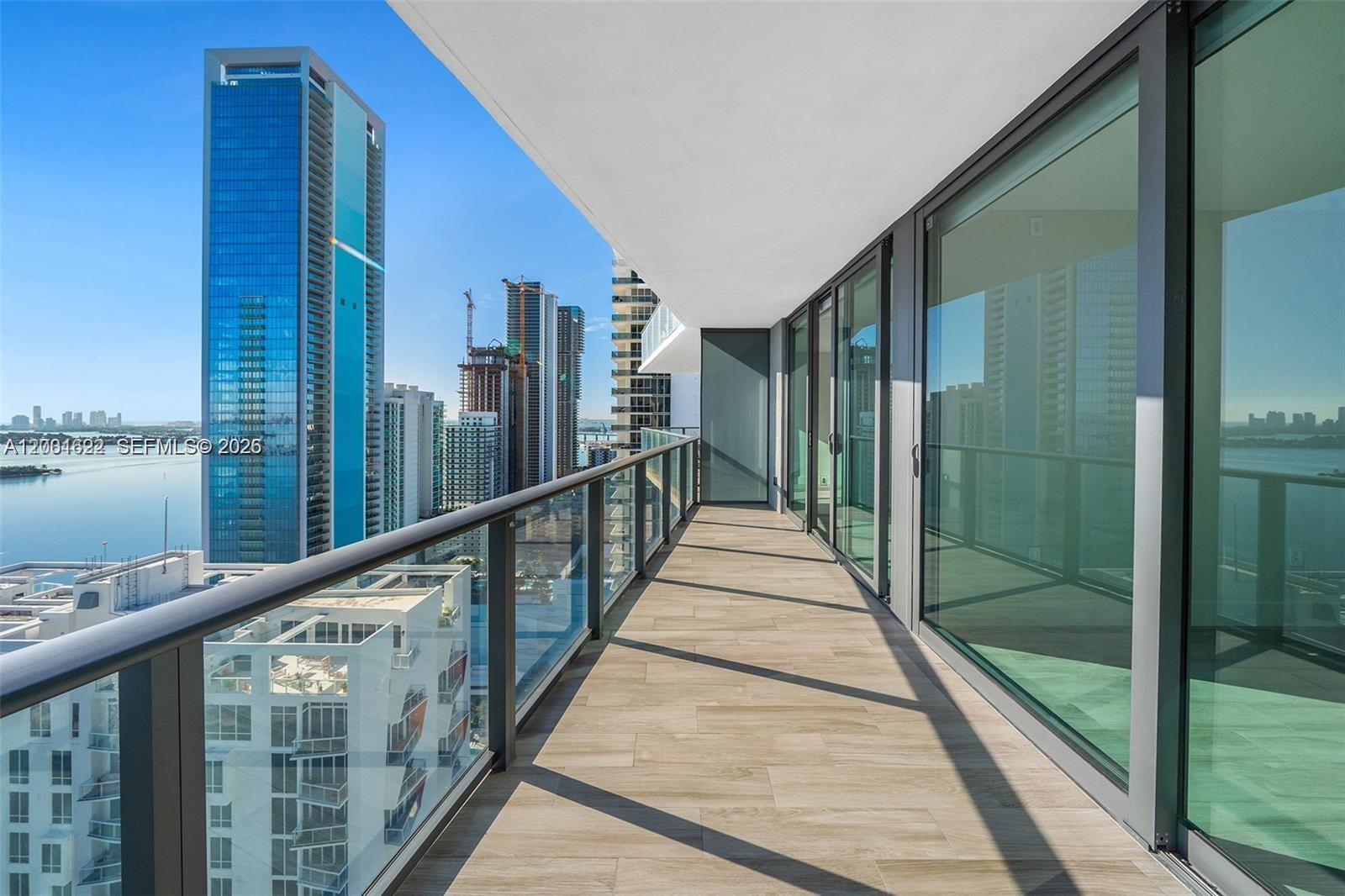 A high-rise balcony features wood-look tile flooring, glass railings, and expansive city and water views — 460 NE 28th St # 2707, Miami FL 33137, MIAMI, FL