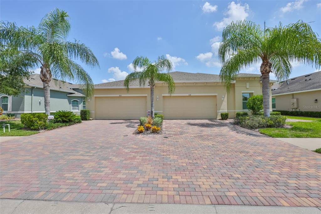 A beige duplex with two garage doors and palm trees sits behind a brick driveway — 606 CHIPPER DRIVE, SUN CITY CENTER, FL