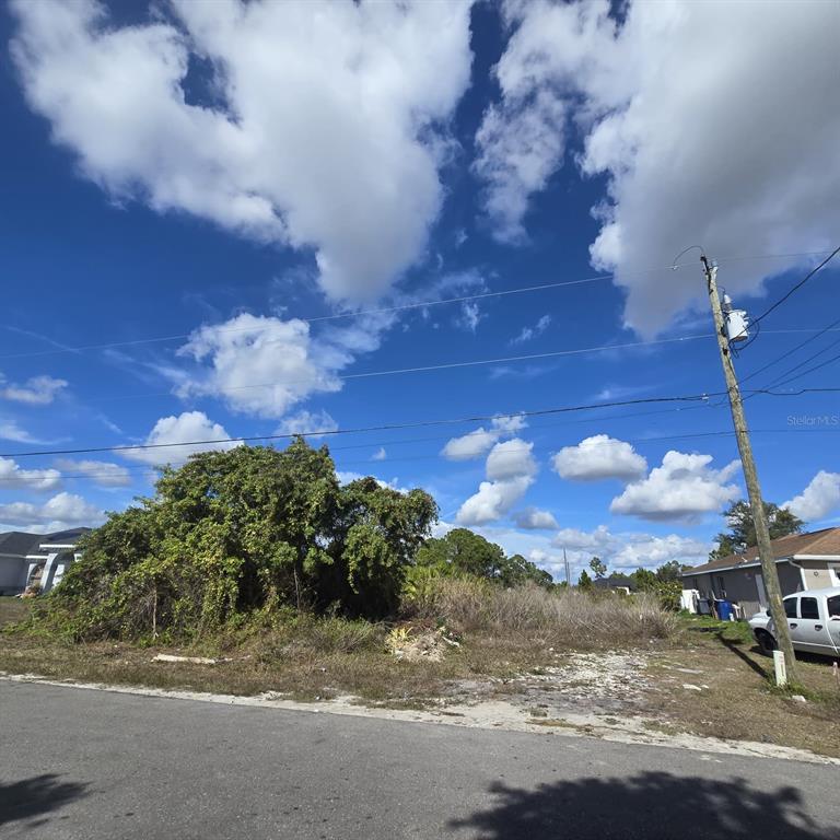 A vacant, overgrown residential lot sits between two houses under a bright, cloudy blue sky — 2804 E 11TH STREET, LEHIGH ACRES, FL