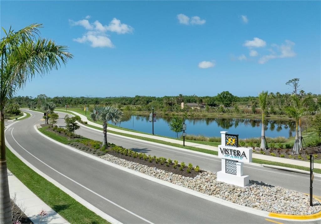 A white community entrance sign stands beside a curved road and a large pond — 332 VISTERA BOULEVARD, NOKOMIS, FL