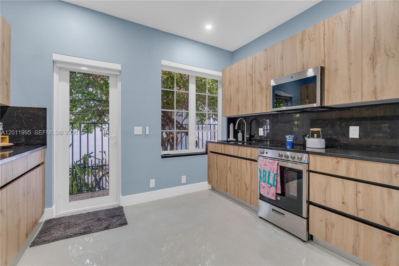 A kitchen features light wood cabinets, black countertops, stainless steel appliances, and a glass door — 1569 Euclid Ave # 7, Miami Beach FL 33139, MIAMI BEACH, FL