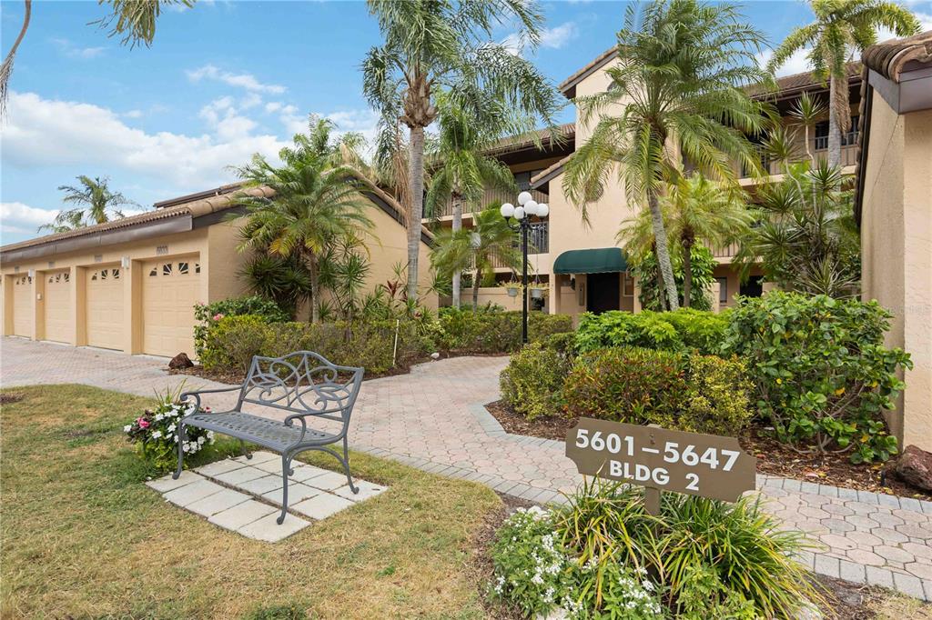 A metal bench sits on a paved walkway in front of a multi-story apartment building — 5627 ASHTON WAY UNIT 5627, SARASOTA, FL