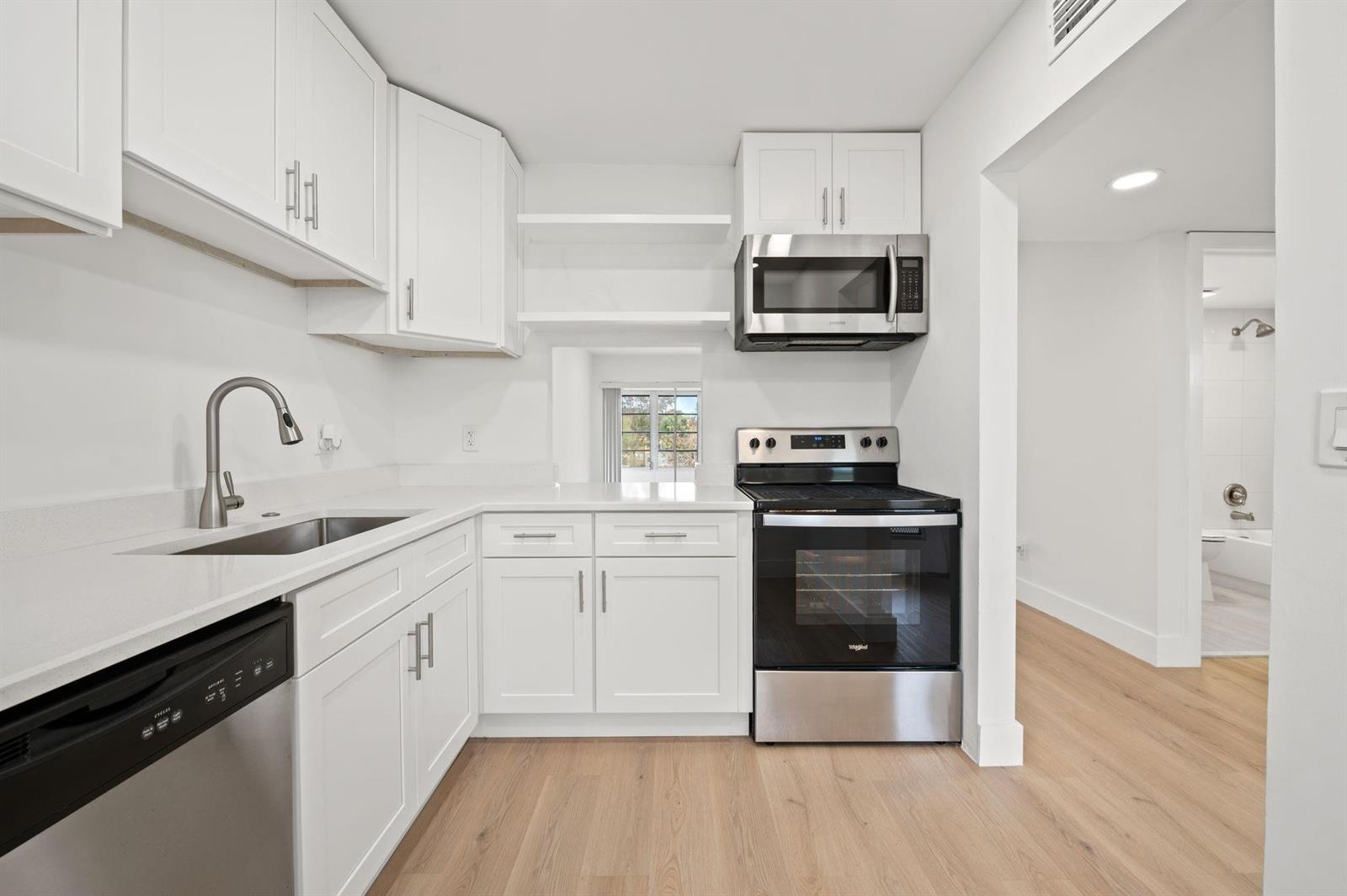 A modern kitchen features white cabinets, stainless steel appliances, and light wood-look flooring — 6000 NW 64th Avenue # 303, Tamarac FL 33319, TAMARAC, FL