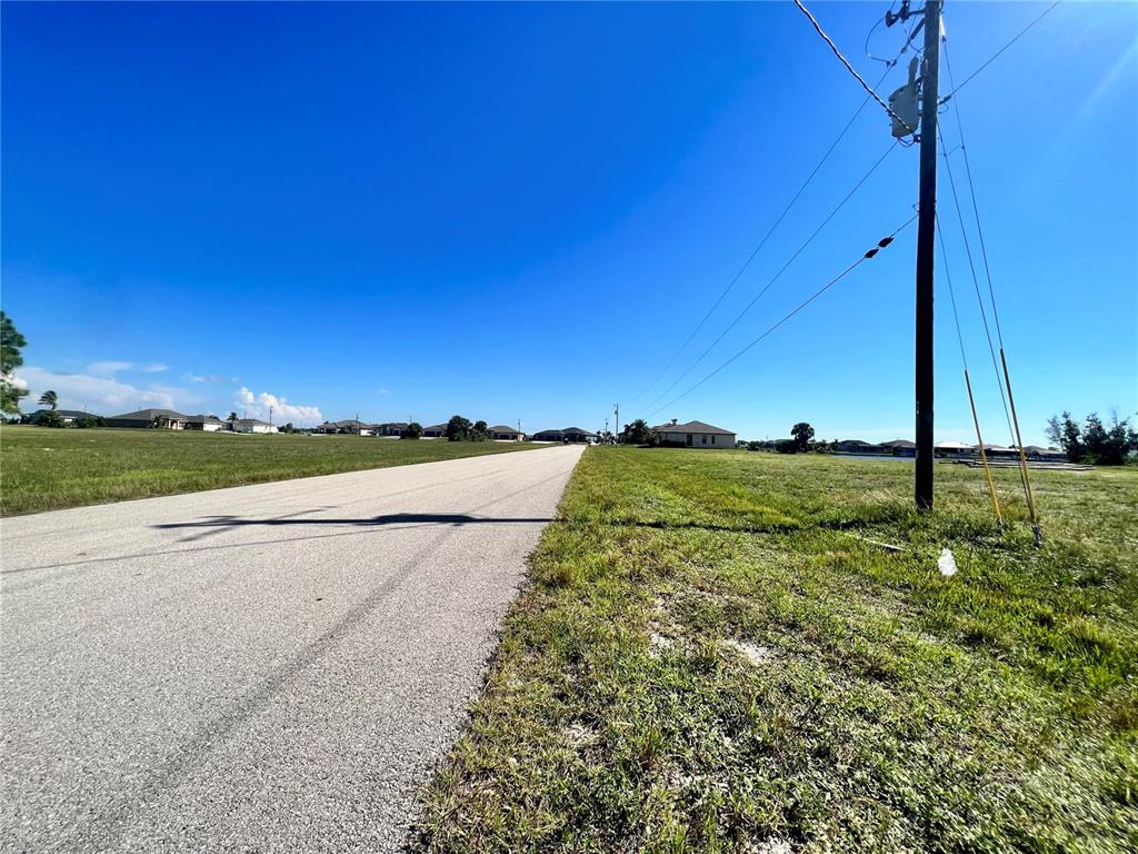 A paved road stretches through a grassy, undeveloped residential area under a clear blue sky — 4008 NW 42ND PLACE, CAPE CORAL, FL