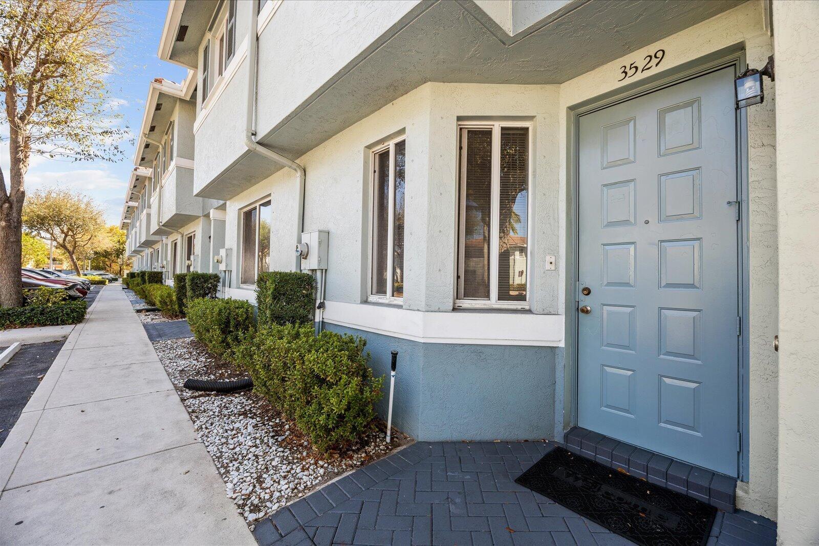 A light blue townhouse exterior features a front door, bay windows, and a paved walkway — 3529 Sonoma Drive, Riviera Beach FL 33404, RIVIERA BEACH, FL