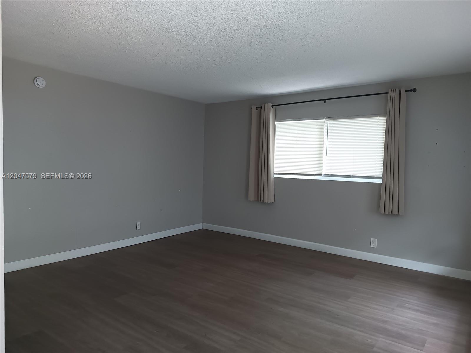 An empty room features gray walls, dark wood-look flooring, and a window with beige curtains — FORT LAUDERDALE, FL