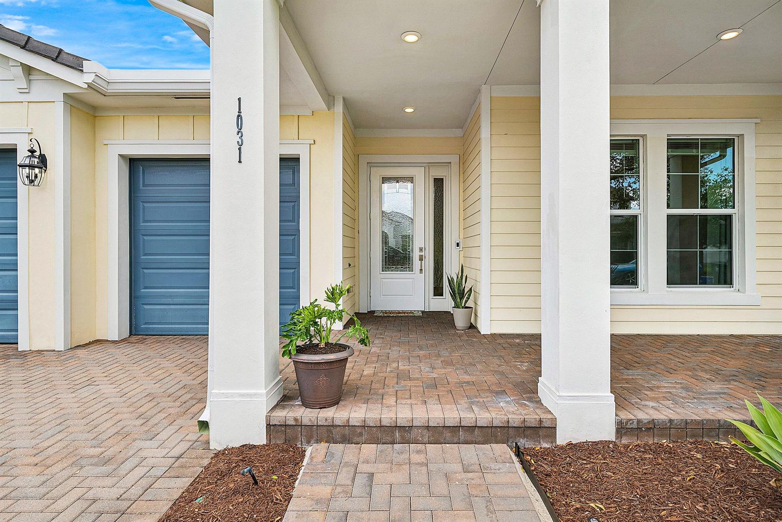 The exterior of a house features a brick walkway, a covered porch, and blue garage doors — 1031 Ember Ridge Run, Loxahatchee FL 33470, LOXAHATCHEE, FL