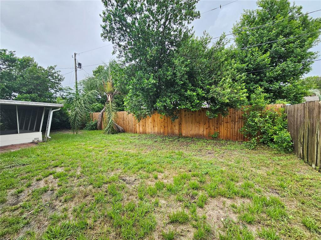 A grassy backyard features a wooden privacy fence, mature trees, and a screened-in patio area — 1702 AQUA LANE, HOLIDAY, FL
