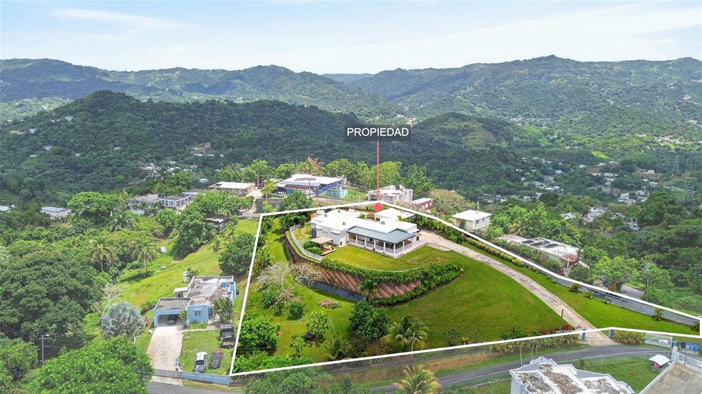 An aerial view shows a white house situated on a grassy hillside surrounded by mountains — PR-839 LOT 9, KM 6.7 ALTOS DE CAMARONES, GUAYNABO, PR