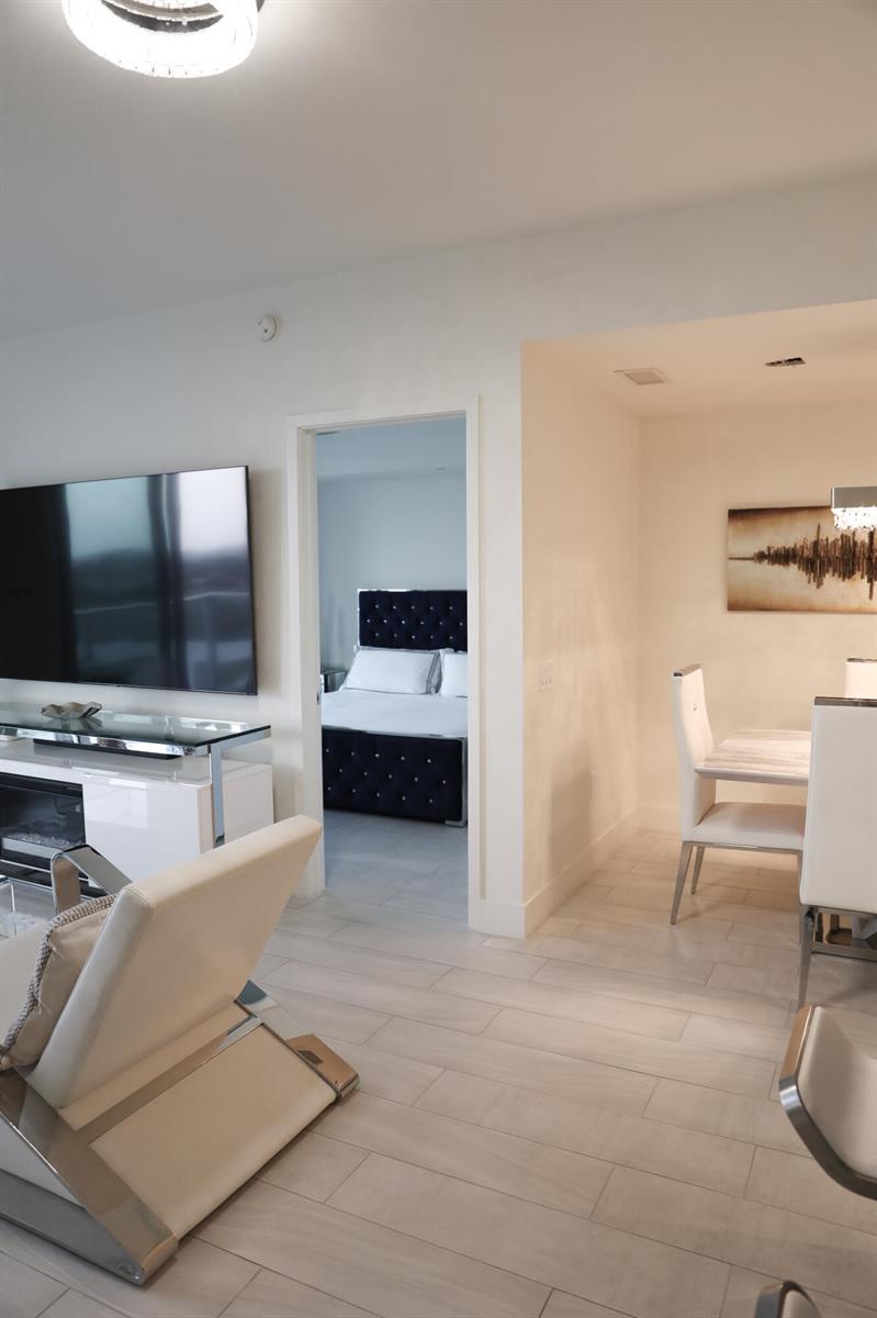 A living room with a television and white furniture leads into a bedroom and dining area — 2000 Metropica Way # 811, Sunrise FL 33323, SUNRISE, FL