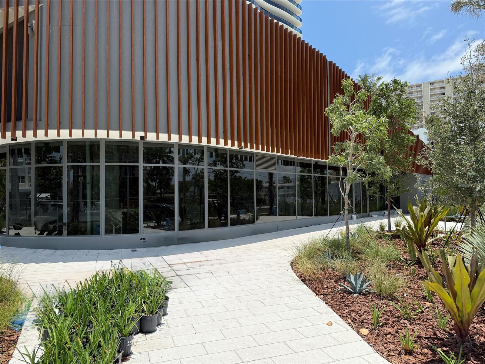 A curved building exterior features vertical wood-look slats above a glass-walled ground floor and landscaping — 900 N Ocean Boulevard, Pompano Beach FL 33062, POMPANO BEACH, FL