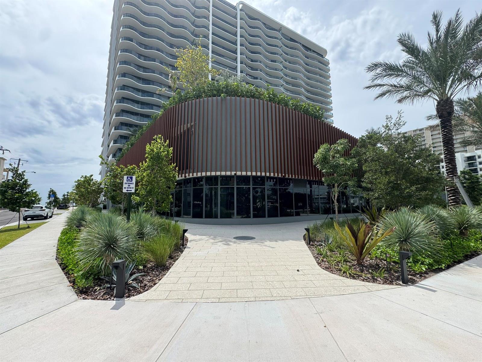 A modern high-rise building features a curved, slatted facade and a landscaped pedestrian walkway — 900 N Ocean Boulevard, Pompano Beach FL 33062, POMPANO BEACH, FL