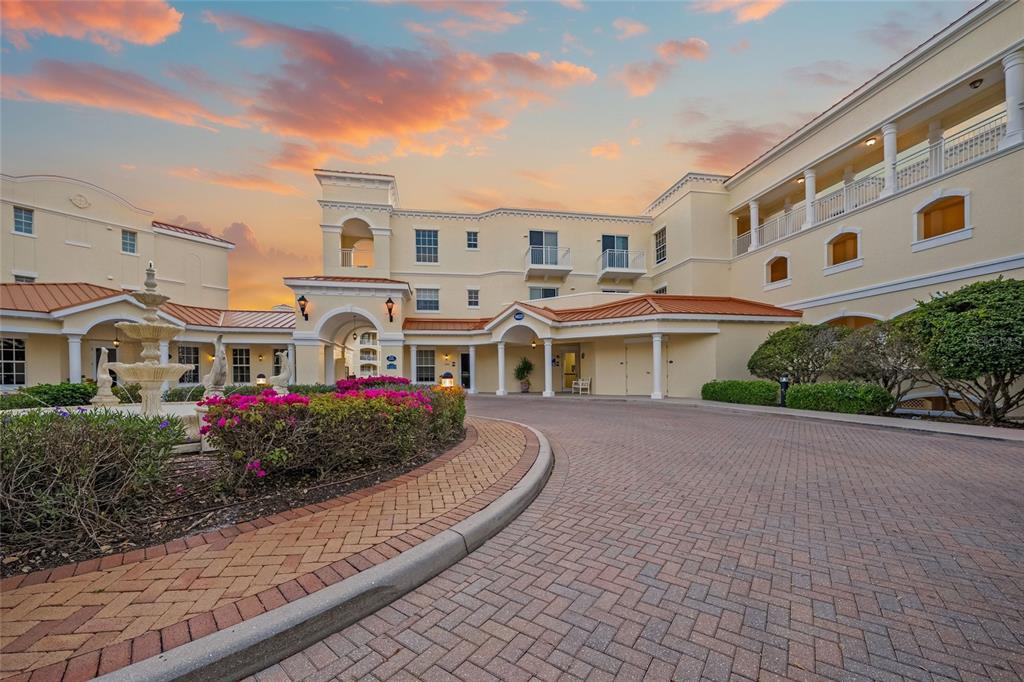 A multi-story building with a brick driveway, a fountain, and pink flowering bushes at sunset — 14021 BELLAGIO WAY UNIT 309, OSPREY, FL