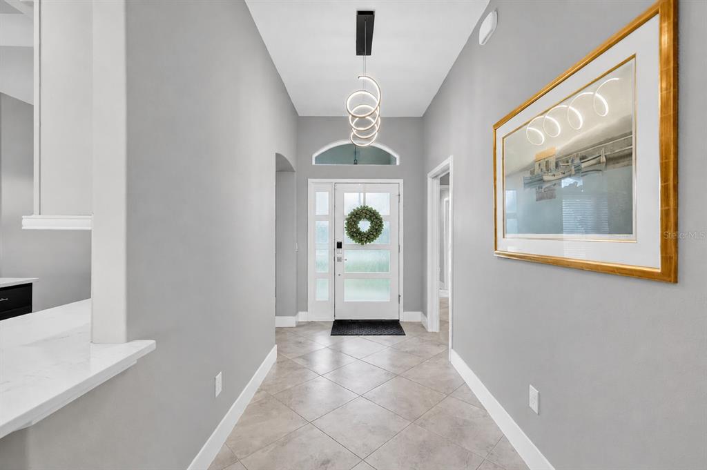 A bright entryway features gray walls, tile flooring, a modern chandelier, and a front door — 2165 GORDON PATH, THE VILLAGES, FL